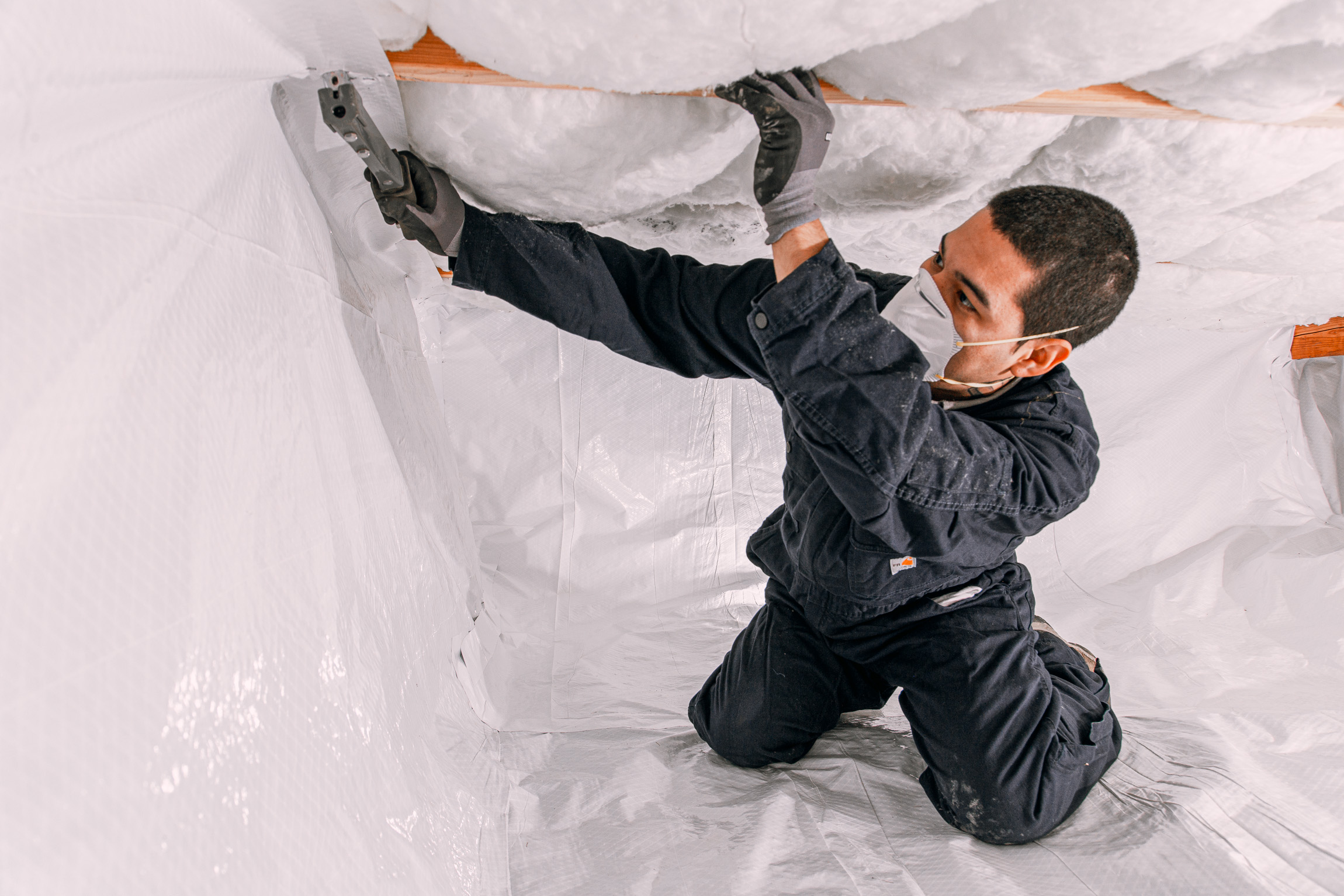 Technician wearing protective gear securing insulation in a crawl space using a fastening tool in Lacey, WA.