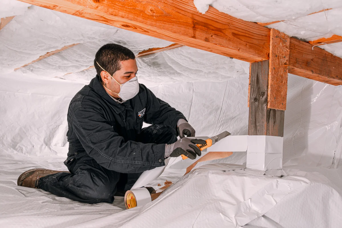 Technician in protective gear sealing a crawl space support beam with insulation tape in Lacey, WA.